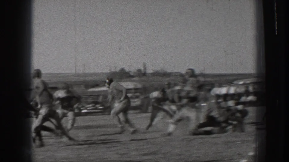 Handheld 8mm frame from the football scrimmage showing players running and blocking across the field, with motion blur, cars, and spectators visible in the distance.
