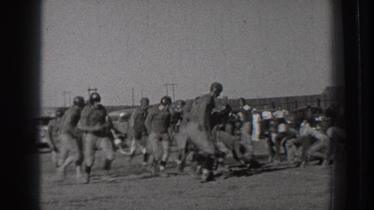 Football players in uniforms gather and move on an open field with spectators and cars behind them.