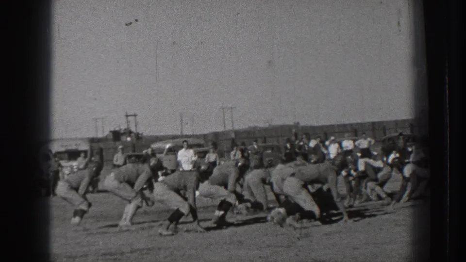 Men in football uniforms line up across the field in a 1939 Washington home-movie view, with onlookers and cars beyond the play.