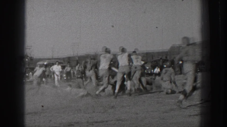 Uniformed players continue a football play across an open Washington field in 1939, with sideline spectators and parked cars in view.