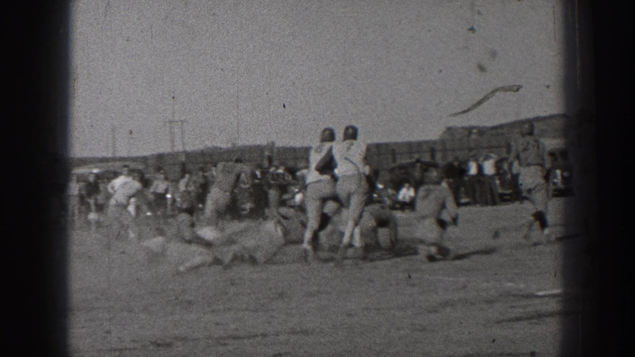 Football players in light and dark uniforms cluster near a tackle with spectators behind them.