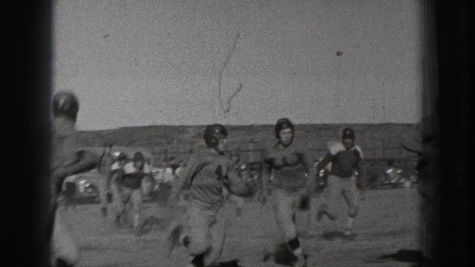 Young male football players in 1939 Washington appear to run on an open field with fencing and hills behind them.