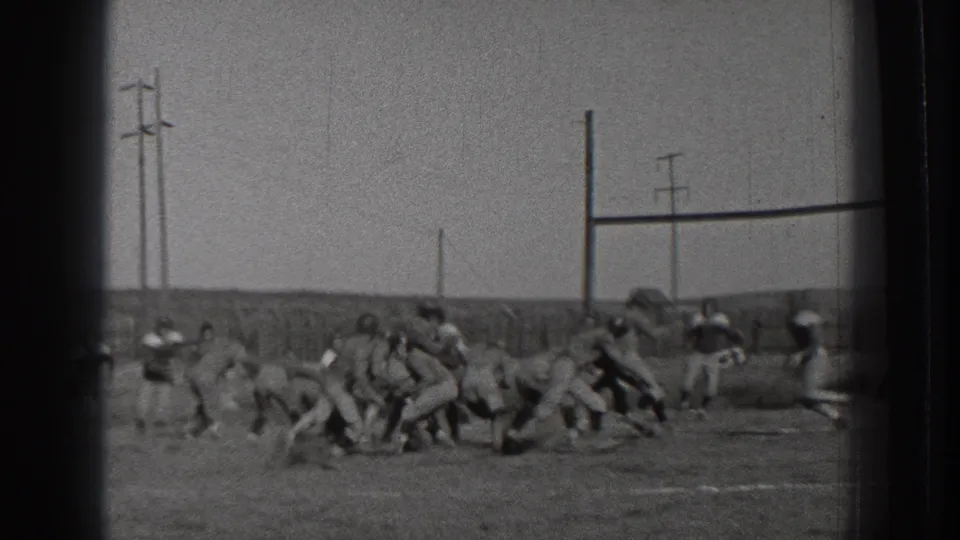 Young male football players in 1939 Washington push and run through a play on an open field bordered by fencing and poles.