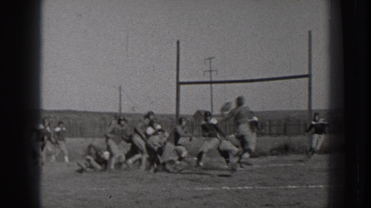 Football players run and tackle near simple goalposts on an outdoor field.