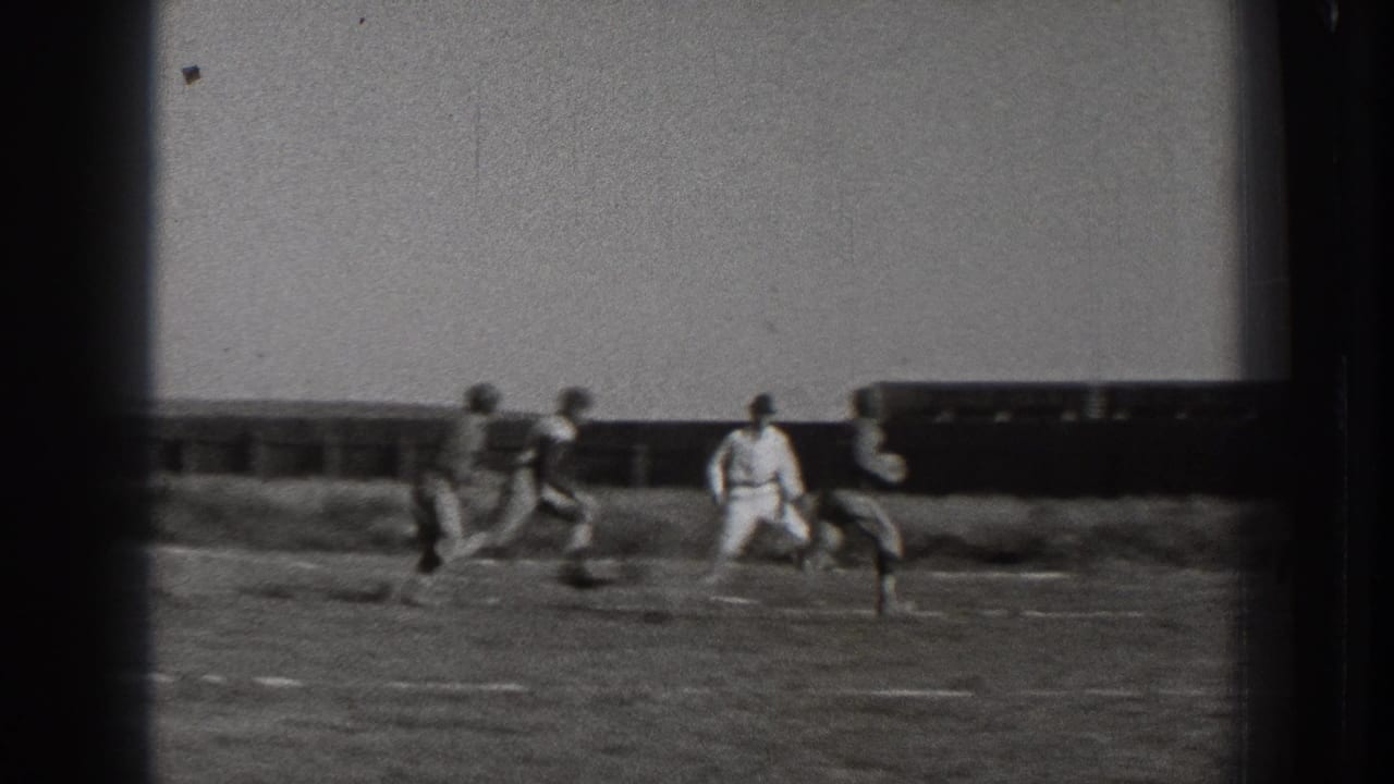 Players run across an outdoor football field with a dark vehicle or structure in the background.