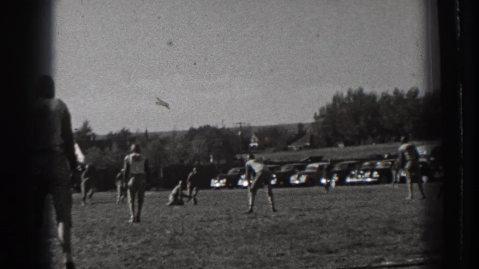 1939 Washington 8mm frame showing the football field from the sideline with players, parked classic cars, trees, and distant buildings.