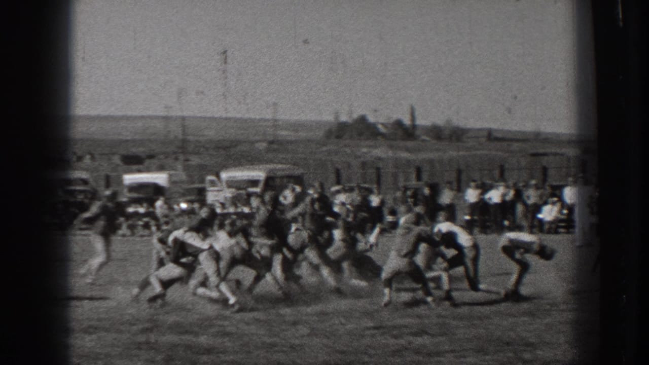 Football players collide on an open field as spectators and cars line the far sideline.