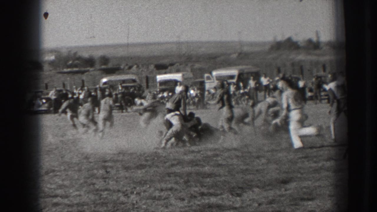 Football players run across a dusty field with spectators and vintage cars behind them.