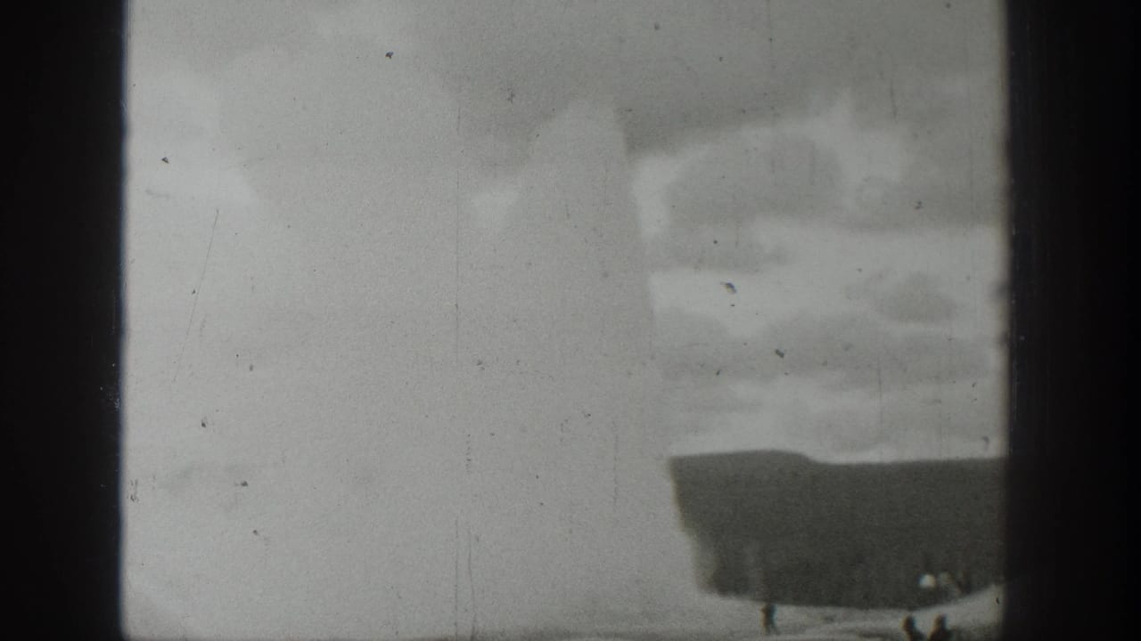 1939 Wyoming 8mm frame showing a pale geyser plume, dark basin edge and small distant spectators under a cloudy sky.