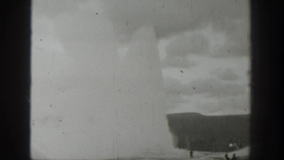 1939 Wyoming 8mm frame showing a pale geyser plume, dark basin edge and small distant spectators under a cloudy sky.