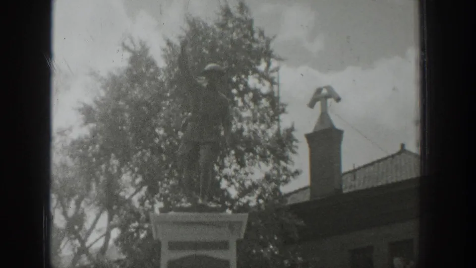 1939 Wyoming home-movie frame with a soldier memorial statue, leafy trees, and a building roofline in soft monochrome.