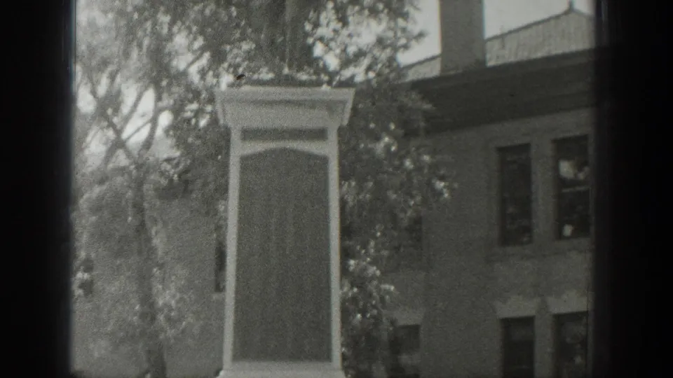 1939 Wyoming frame showing the memorial pedestal beside trees, with a masonry building and windows in the background.