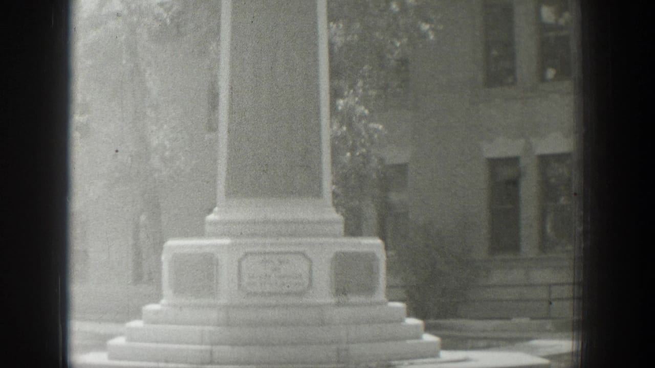 Base of a stone memorial with steps, plaques, trees, and a building behind it.