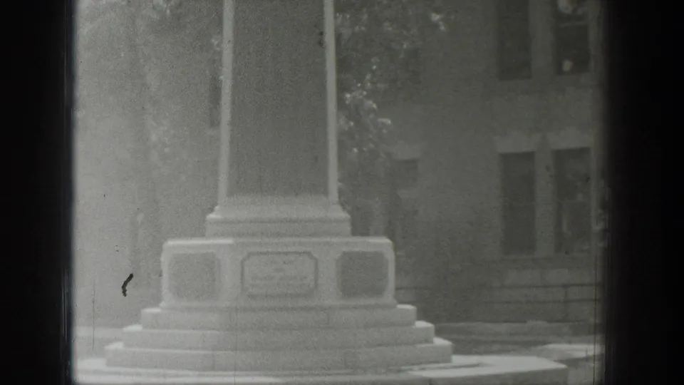 1939 Wyoming 8mm frame showing the lower memorial column and base plaques, with a building partly visible behind foliage.