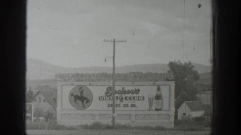 1939 Wyoming 8mm frame showing a roadside dining club and beer billboard with a cowboy logo, bottle graphic, and hills behind it.
