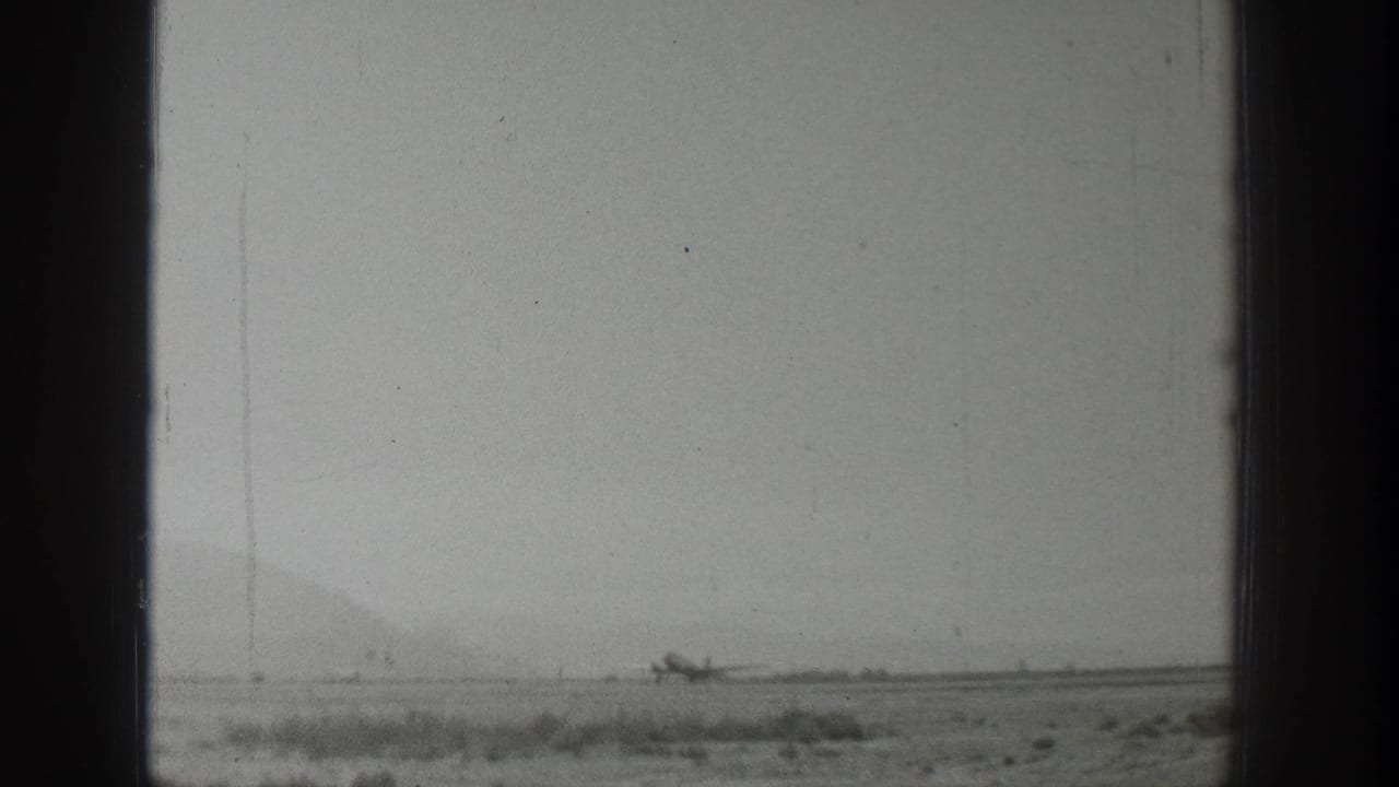1939 Wyoming 8mm frame showing a small propeller airplane on a flat dirt runway, seen through dark framing with hazy mountains beyond.