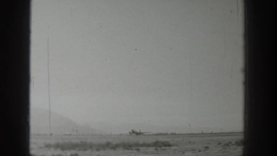 1939 Wyoming 8mm frame showing a small propeller airplane on a flat dirt runway, seen through dark framing with hazy mountains beyond.