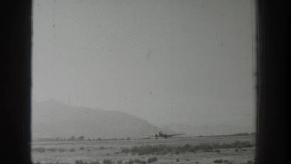 Small airplane taxis across a remote Wyoming runway in 1939, with scrubland foreground, hazy mountains, and dark window framing.