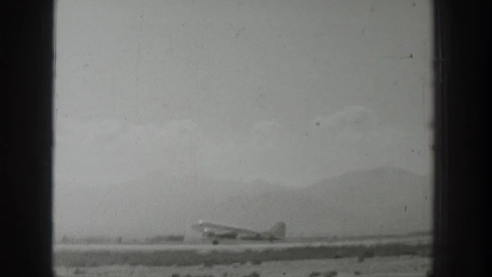 Black-and-white 1939 Wyoming 8mm frame showing a propeller airplane on a runway before distant mountains.