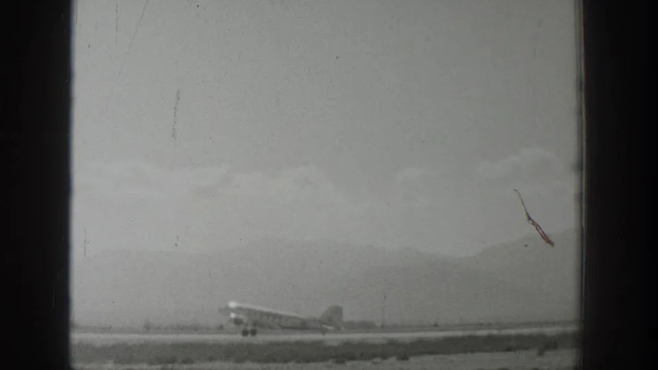 Black-and-white 1939 Wyoming 8mm frame of a small propeller airplane progressing along a runway with mountains behind.
