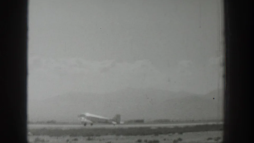 Black-and-white 1939 Wyoming 8mm frame showing the airplane lower in the frame on an open runway with hills beyond.
