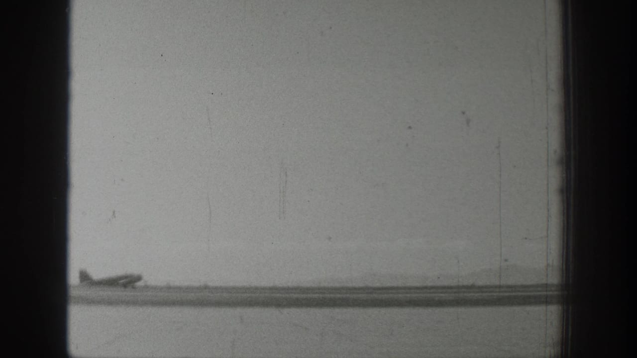 Small propeller airplane on a flat strip near water, with distant hills and film scratches visible.