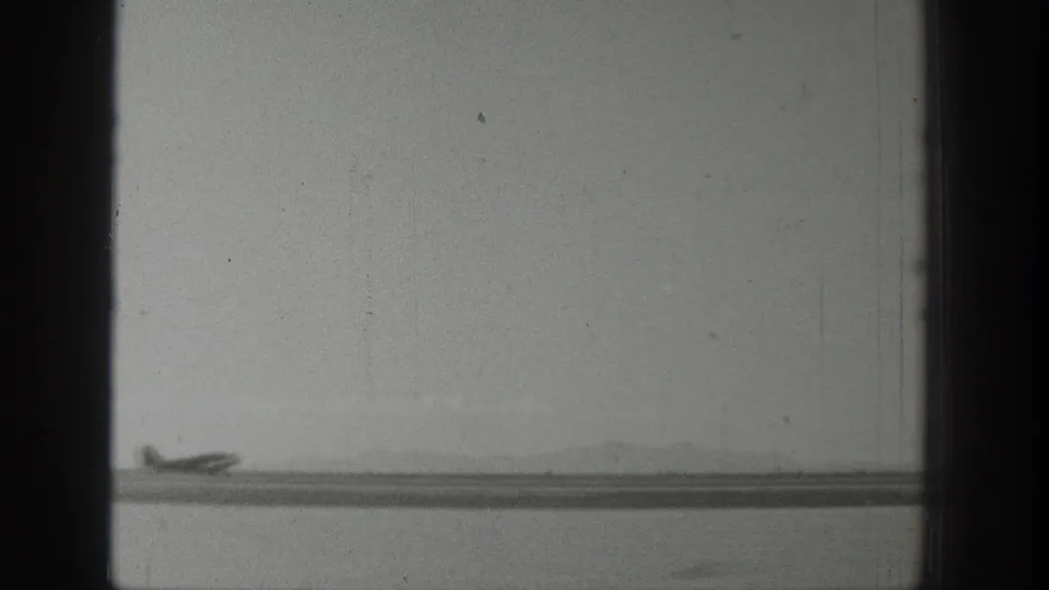 Wyoming 1939 home-movie frame with a small propeller airplane near the left edge of a broad riverside strip and distant hills.
