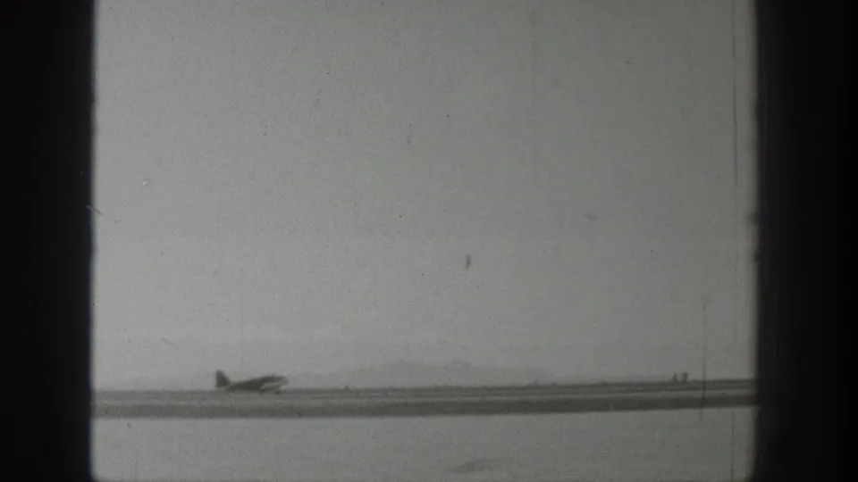 Wyoming 1939 frame showing a small airplane on the ground near a river or shoreline, with low mountains in the background.
