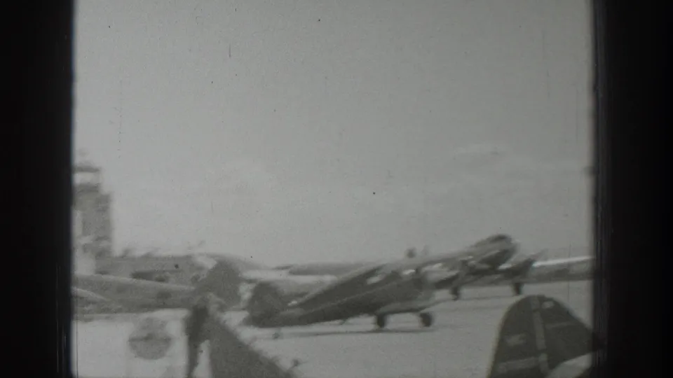 1939 Wyoming 8mm frame with multiple propeller aircraft near a runway and a small tower-like airfield structure in the background.