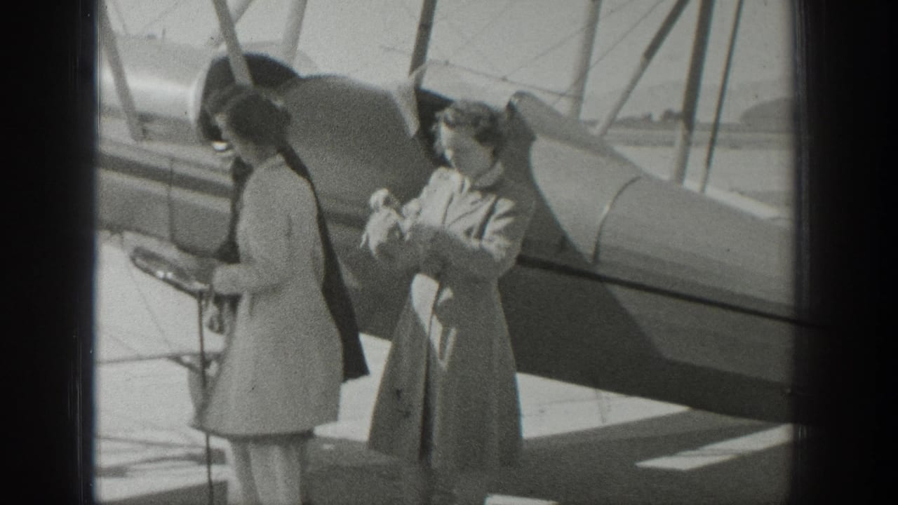 Two women in coats and hats stand beside a biplane on an airfield.