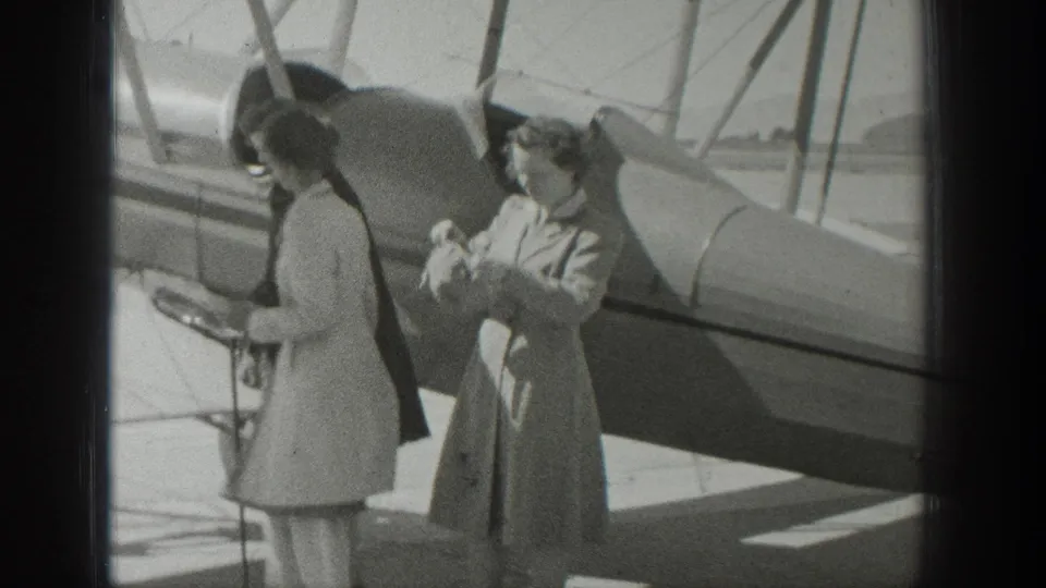 1939 Wyoming home-movie frame showing two women near a biplane, with the fuselage and wing struts visible behind them.