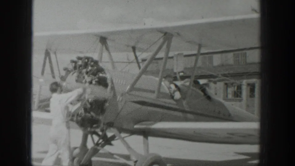 1939 Wyoming frame showing the biplane from the side, with a person by the engine, the open cockpit, wing struts, and landing gear visible.
