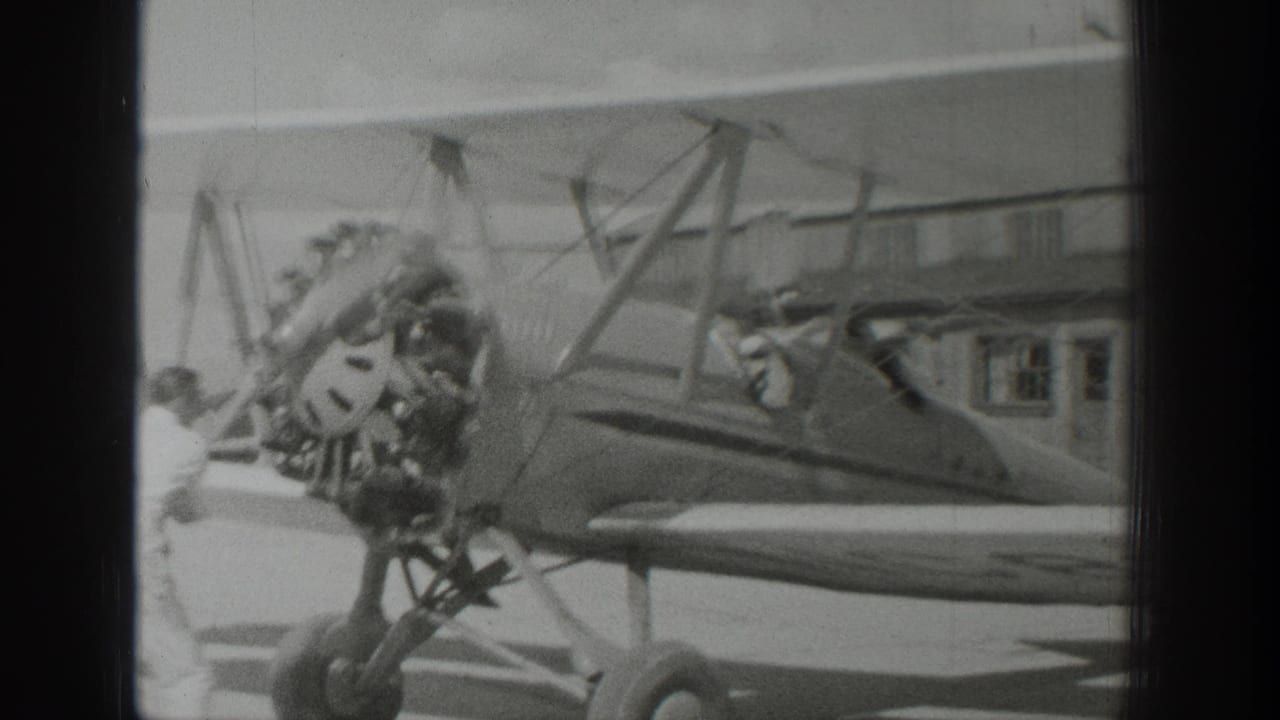 1939 Wyoming home-movie frame showing a parked biplane near a hangar, with radial engine, propeller, struts, and open cockpit visible.
