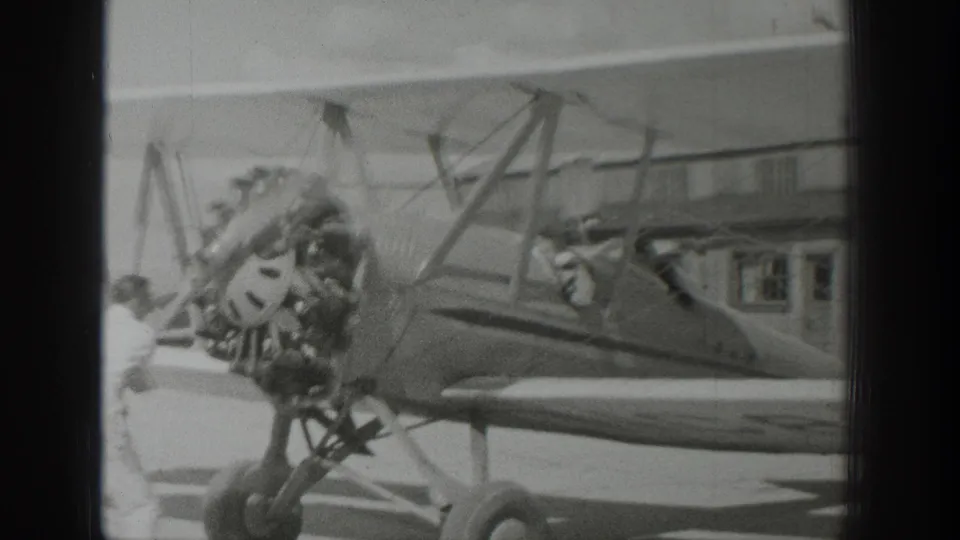 1939 Wyoming home-movie frame showing a parked biplane near a hangar, with radial engine, propeller, struts, and open cockpit visible.