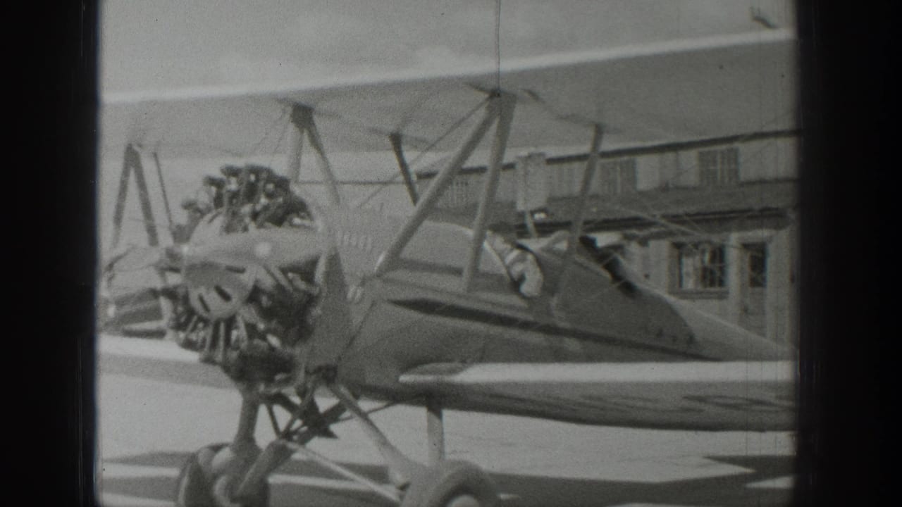 Side view of the biplane with propeller, radial engine, struts, cockpit, and hangar behind it.