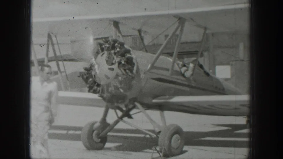 A man in light clothing stands near the biplane's radial engine and landing gear in this 1939 Wyoming airfield frame.