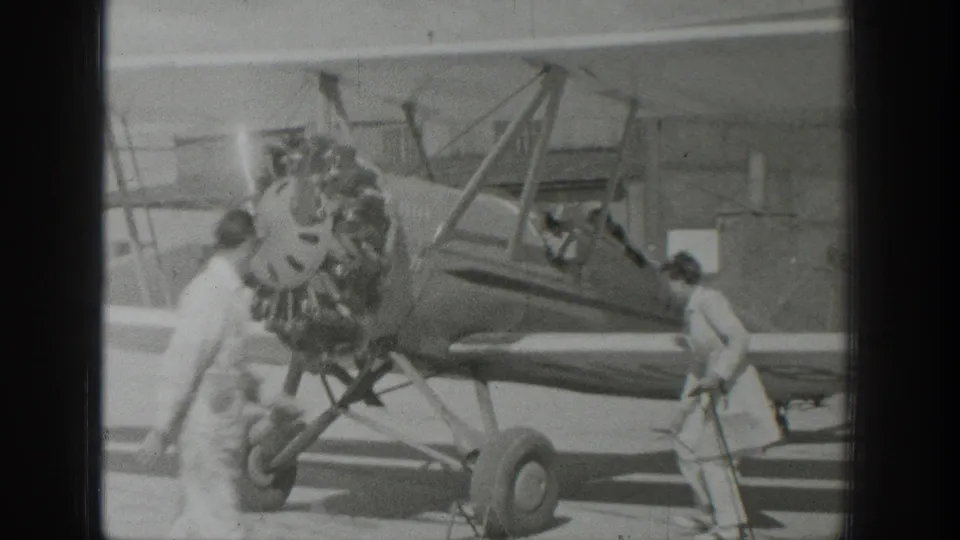 Two people are visible near the parked biplane in 1939 Wyoming footage, with a bright flare near the radial engine and the hangar behind.