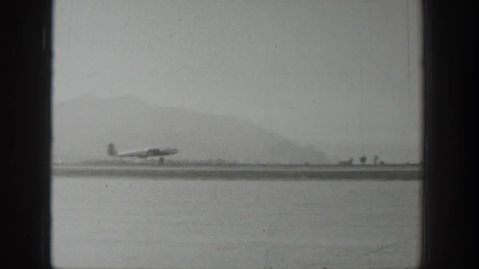 Wyoming 1939 home-movie frame showing a small airplane on or near touchdown on a distant airstrip with hazy mountains beyond.