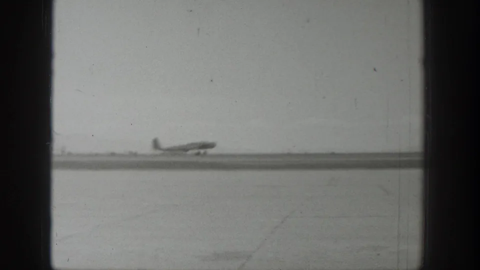A small propeller aircraft appears farther down the Wyoming runway, framed by grainy black-and-white 8mm film edges.