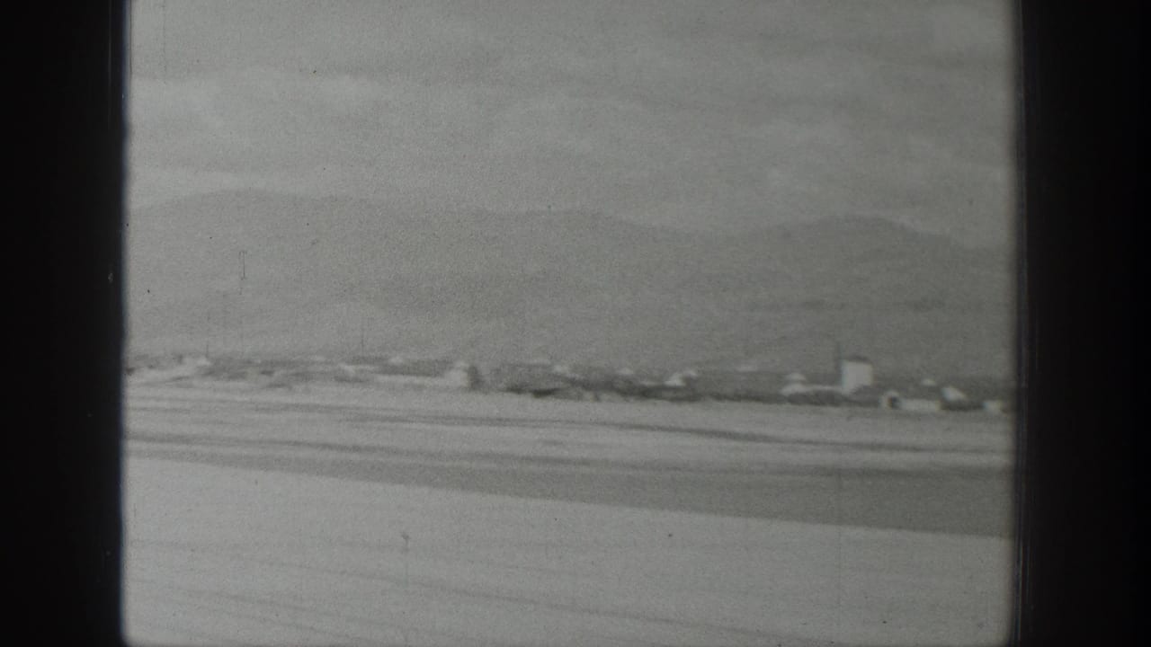 Black-and-white aerial home-movie frame over Wyoming, with scattered structures, pale ground, and low mountains seen past the plane window.