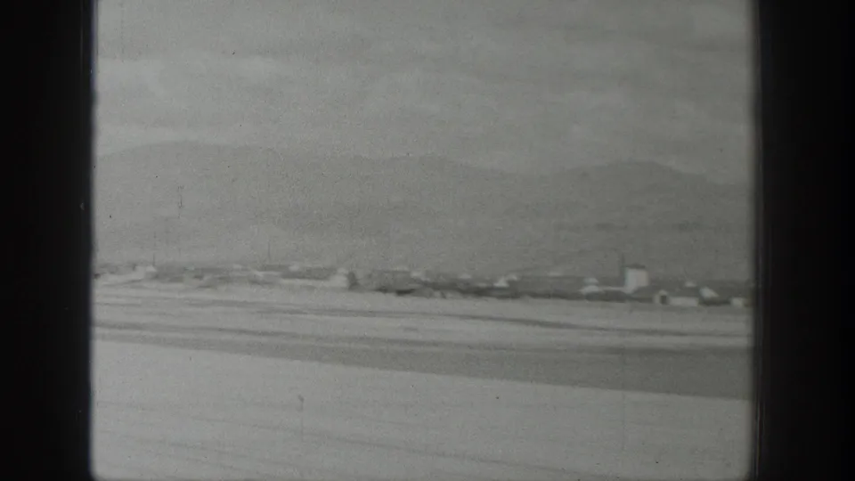 Black-and-white aerial home-movie frame over Wyoming, with scattered structures, pale ground, and low mountains seen past the plane window.