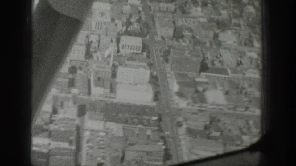 Black-and-white 8mm aerial view through glass, showing rooftops, streets, and a curved aircraft structure at the edge.