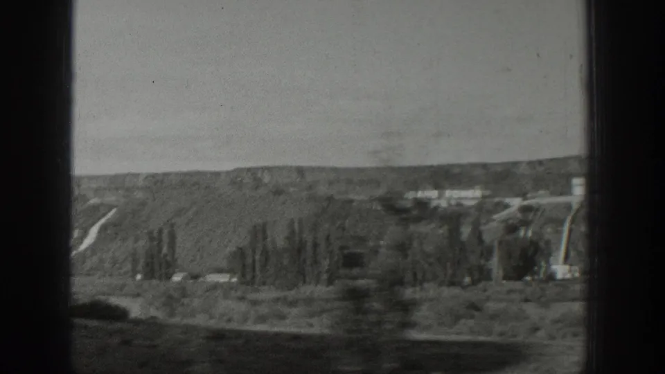 The moving 8mm view looks across a dry Wyoming hillside toward distant buildings, with dark window bars cutting through the frame.
