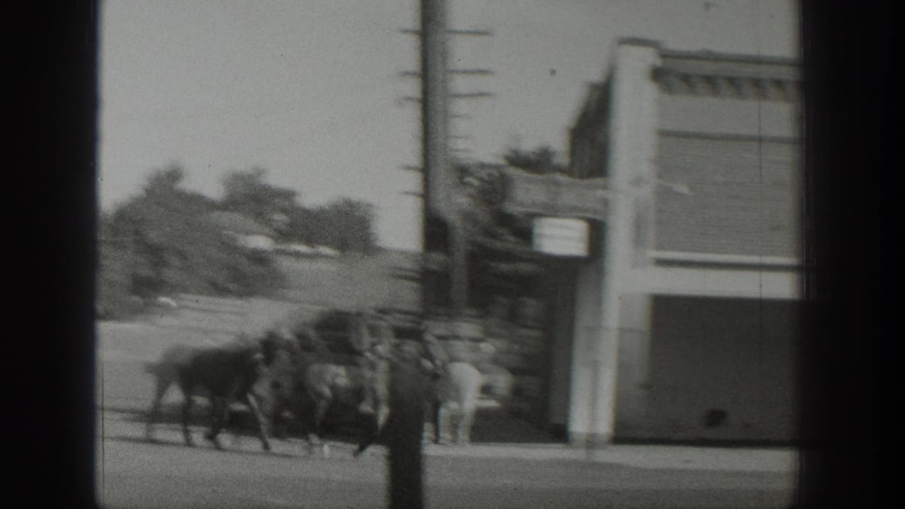 Horses move past a corner storefront and utility pole in the supplied 1939 Wyoming home-movie scene, with riders and street buildings visible.