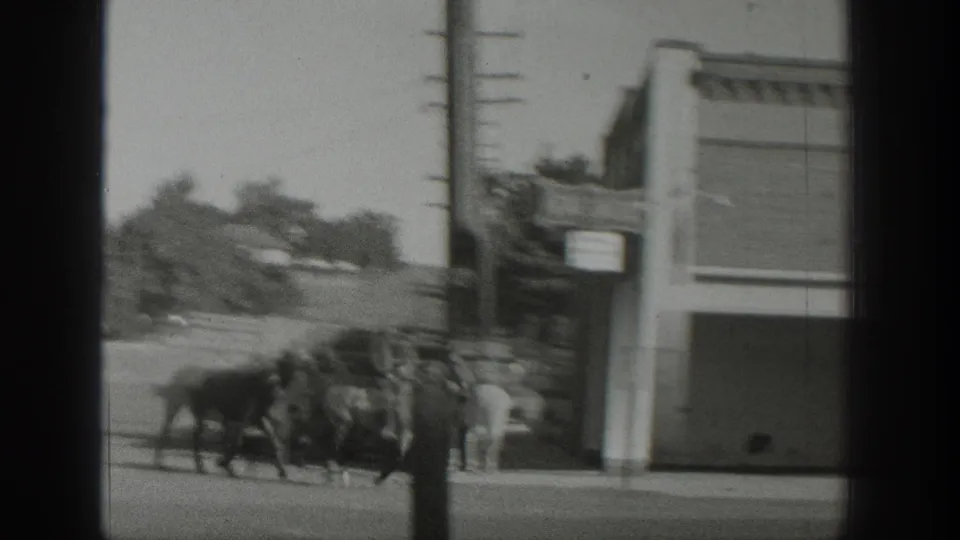Horses move past a corner storefront and utility pole in the supplied 1939 Wyoming home-movie scene, with riders and street buildings visible.