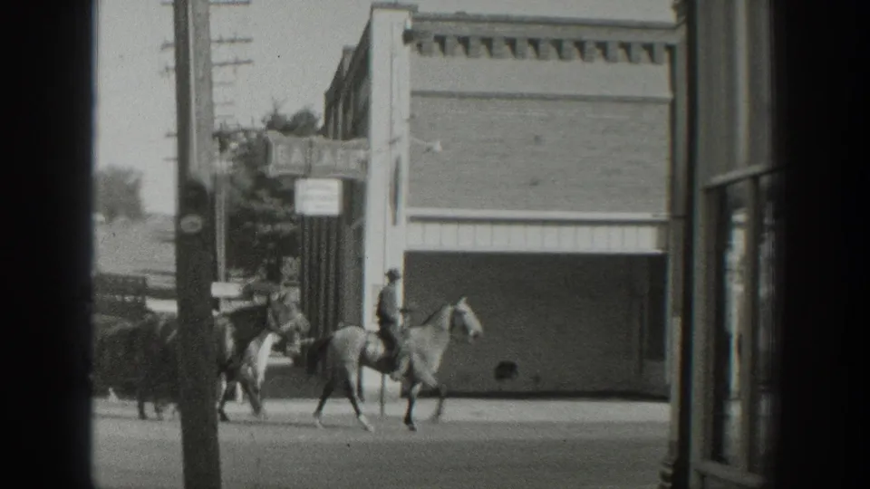 A mounted rider on a light-colored horse leads other horses along the street in Wyoming, with a commercial facade and pole in view.