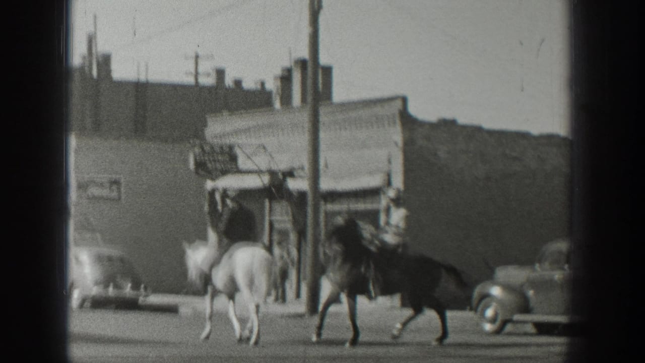 In 1939 Wyoming home-movie footage, two riders on horseback move along a street past storefronts, utility poles, and rounded cars.