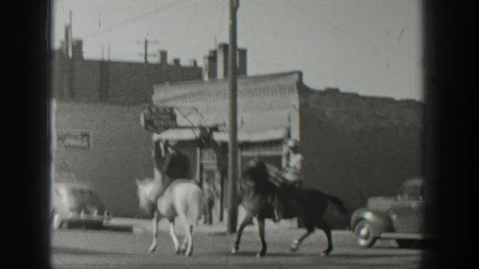 In 1939 Wyoming home-movie footage, two riders on horseback move along a street past storefronts, utility poles, and rounded cars.