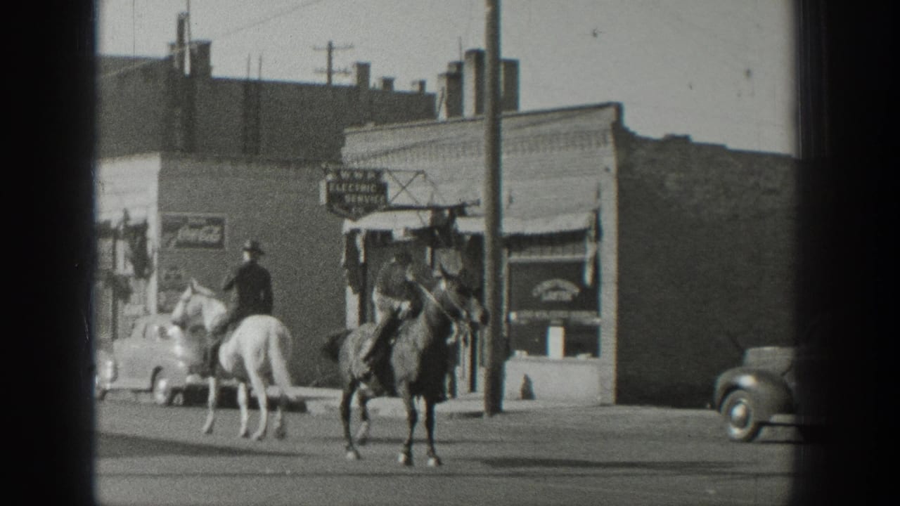 Two cowboy-hatted riders on a white and dark horse pass storefront signage and rounded automobiles in 1939 Wyoming footage.