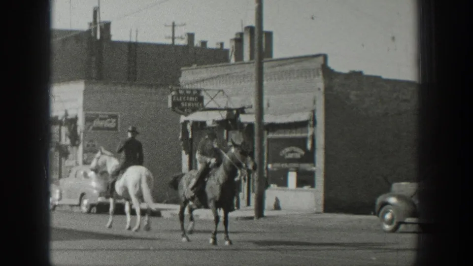 Two cowboy-hatted riders on a white and dark horse pass storefront signage and rounded automobiles in 1939 Wyoming footage.